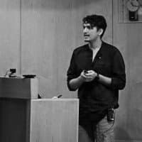 A man standing at a podium or lectern, holding a device, possibly a phone or remote, in a room with wood-paneled walls
